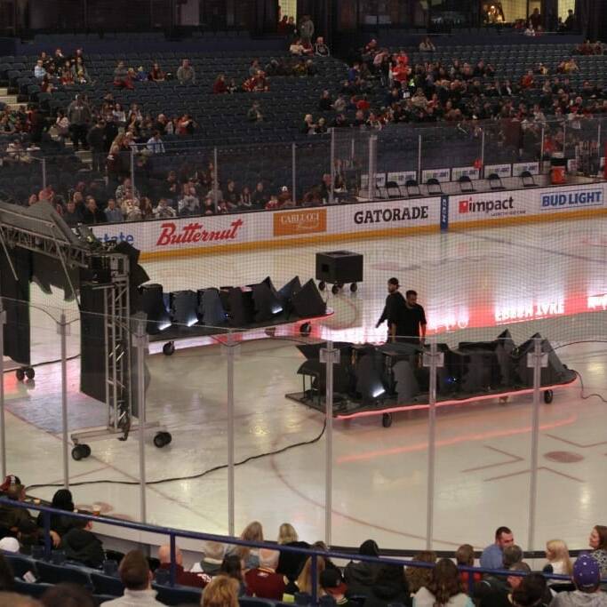 Workers set up lighting equipment on an ice rink before a Chicago Wolves hockey game. The rink is surrounded by advertising boards for various brands, and spectators are seated in the stands, eagerly awaiting the 2018 March Networking Night. The arena's atmosphere is lively with anticipation.