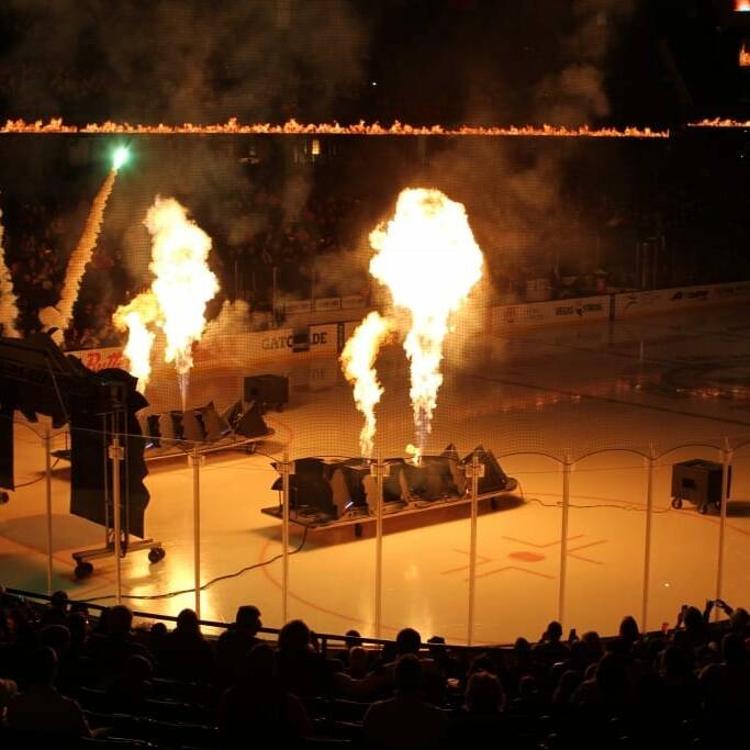 A hockey arena is illuminated by large flames shooting up from multiple pyrotechnic devices on the ice, set up for a dramatic pre-game show. Bright lights and fireworks add to the spectacle as the audience at the 2018 Chicago Wolves Networking Night watches from the darkened stands.