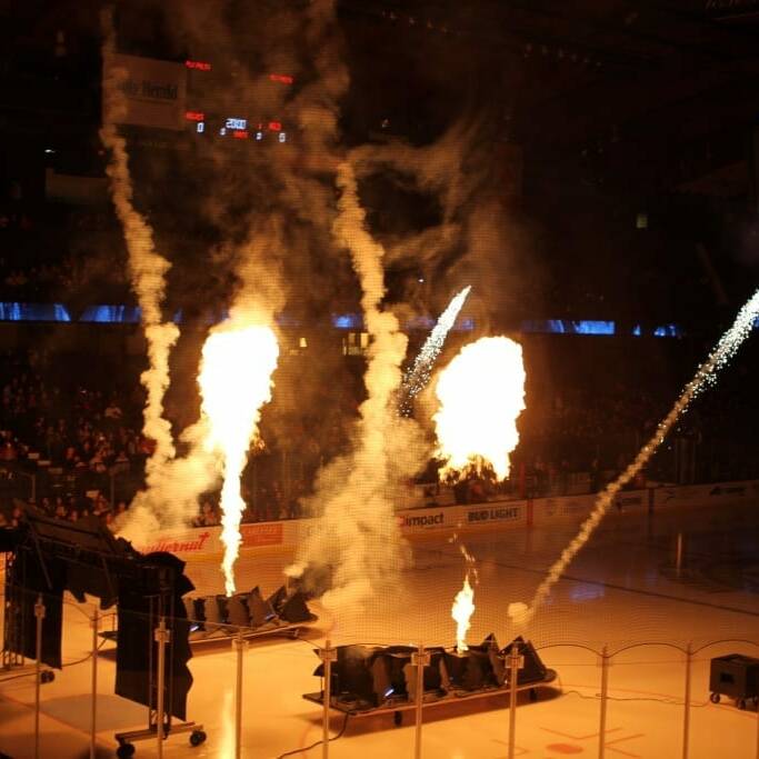 A hockey arena with pyrotechnics shooting flames and sparkles into the air, creating a fiery and dazzling display. During the 2018 March Networking Night, a smoke-filled stage is set up in front of the ice rink, with crowd lights and the Chicago Wolves' large screen in the background.