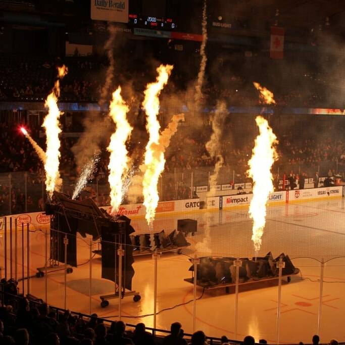 An indoor ice hockey arena with pyrotechnic flames shooting up near the seating area creates a dramatic effect, likely during a Chicago Wolves game. The illuminated ice rink is packed with spectators, perhaps part of a special 2018 March Networking Night event, eagerly watching the action unfold.