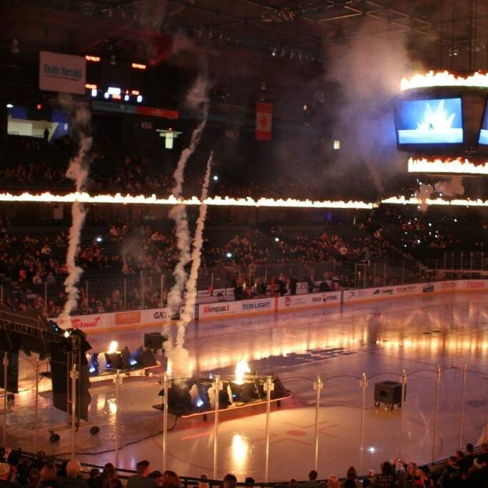 A hockey arena with pyrotechnics going off on the ice. Smoke and flames are rising near the goal area during the Chicago Wolves' 2018 March Networking Night. The grandstands are filled with spectators, and there is a large screen hanging from the ceiling. Bright lights highlight the rink and add to the thrilling atmosphere.