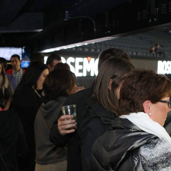 A group of people, some holding drinks, gather in a semi-outdoor setting with bleacher seating visible in the background. The scene suggests an event or gathering, possibly 2018 March Networking Night with Chicago Wolves. Some individuals are engaged in conversation, while others look around.