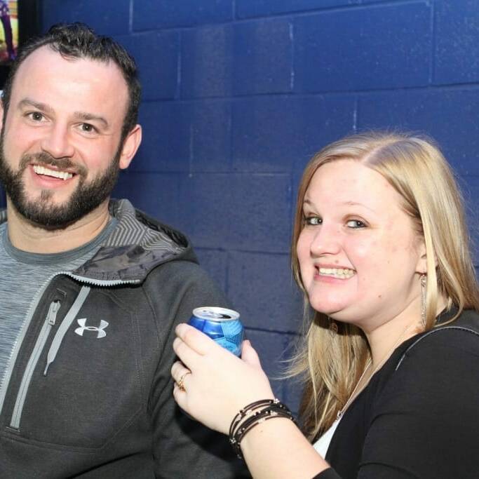 A man and a woman smiling at the camera. The man is wearing a gray Under Armour jacket, and the woman is holding a can of drink and wearing a black top with a white inside layer. They are standing against a blue wall with a partially visible sign in the background, captured during the 2018 March Networking Night hosted by Chicago Wolves.