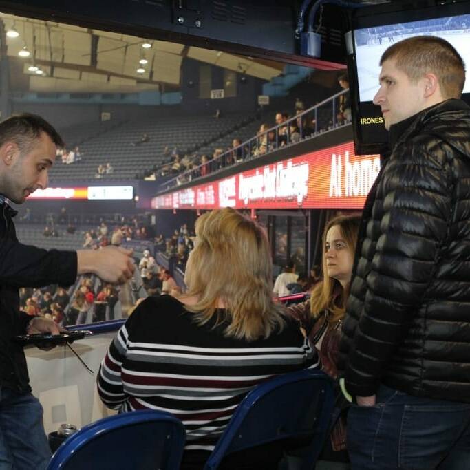 A group of people is standing and sitting in an indoor arena with a hockey game visible in the background. One man is gesturing as he speaks to three others. The arena seats are sparsely filled, and a large digital signboard displays event information for the 2018 March Networking Night with the Chicago Wolves.