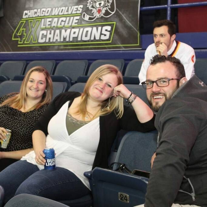 Four people are sitting in stadium seats. Two women and a man are in the front row, smiling and holding canned drinks. Another man is seated behind them. A banner on the wall reads "Chicago Wolves 4X League Champions." It's 2018 March Networking Night at the arena.