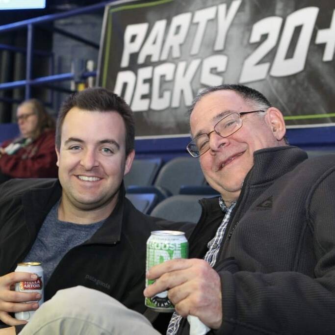 Two men are sitting in stadium seats, smiling at the camera. Both are holding cans of drinks. Behind them is a sign that reads "PARTY DECKS 20+." Other spectators can be seen in the background. The atmosphere appears relaxed and cheerful, typical for a Networking Night at a Chicago Wolves game.