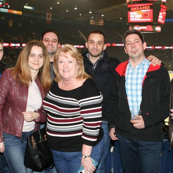 A group of seven people, four men and three women, standing closely together and smiling at the 2018 March Networking Night hosted by the Chicago Wolves. Some hold drinks, dressed in casual attire with jackets. A large crowd and illuminated screens are visible in the background.