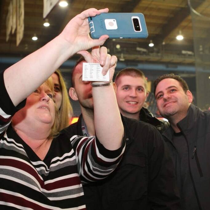 A group of four people is taking a selfie in an indoor stadium setting during the Chicago Wolves Networking Night. The person in front is holding the phone, capturing the moment. All individuals are smiling, and the large indoor setting includes overhead lighting and a scoreboard in the background from 2018.