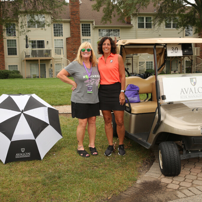 Two women smiling and standing next to a golf cart marked "Avalon" with an umbrella on the grass. They are participating in the 2021 IACAC Golf Outing, with a building in the background. One woman is wearing a grey shirt; the other is in a bright top and black skirt.