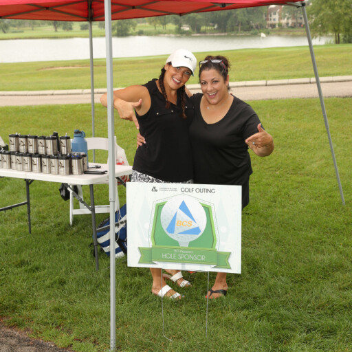 Two women under a red canopy stand next to a sign reading "Hole Sponsor" at the 2021 IACAC Golf Outing. They are smiling and posing for the camera, with one giving a thumbs-up and the other making a peace sign. A lake and various equipment are visible in the background on a cloudy day.