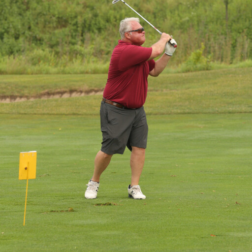 A man with short hair, wearing a red polo shirt, gray shorts, and white sneakers, is playing golf on a green course during the 2021 IACAC tournament. He is mid-swing with a golf club. There is a yellow marker in the grass near him, and trees and a hill are visible in the background.