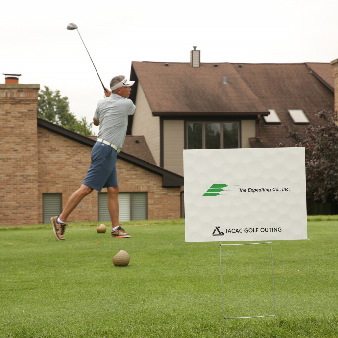 A man is swinging a golf club at a tee on a well-manicured golf course during the 2021 IACAC Golf Outing. Casually dressed and wearing a visor, he seems focused. In the foreground, there's a sign that reads "The Expediting Co., Inc." and "IACAC Golf Outing," with brick buildings and trees in the background.