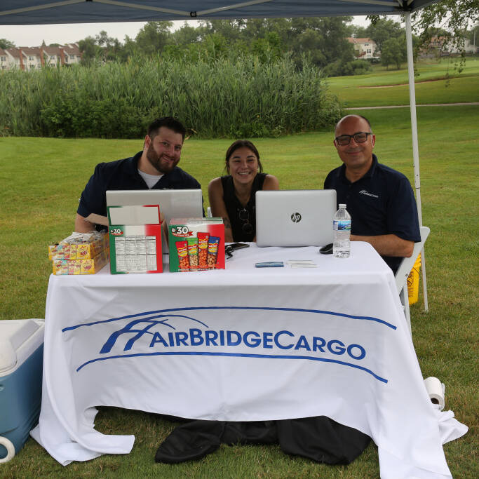 Three people sit at a table under a canopy tent with a sign that reads "AirBridgeCargo" during the 2021 IACAC Golf Outing. The table displays two laptops, snacks, and other items. In the background, there is greenery and residential houses.