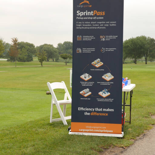 A vertical banner for Cargo Sprint's "SprintPass" system stands on a grassy area by the golf course at the 2021 IACAC Golf Outing. The banner lists features and benefits of the system, with a white folding chair nearby. Trees and a cloudy sky create a picturesque backdrop.