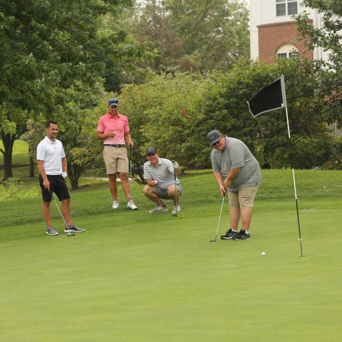 During the 2021 IACAC Golf Outing, four men are on a golf green. One man is putting the ball towards the hole, while the other three watch. The background includes trees and a partial view of a building. The flagstick remains in the hole.