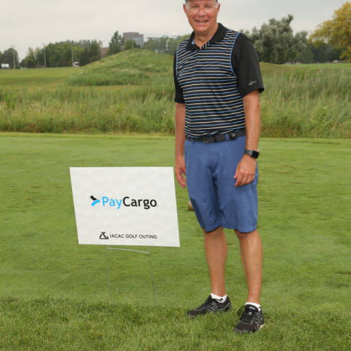 A man wearing a golf hat, black striped polo, and blue shorts stands smiling on a grassy golf course during the 2021 IACAC Golf Outing. He is next to a sign that reads "PayCargo" with a logo and "IACAC Golf Outing" below it. Trees and a cloudy sky are in the background.