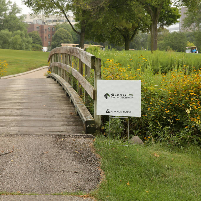 A wooden footbridge leads to a grassy area in the park where the 2021 Golf Outing will be held. To the right of the bridge, an IACAC sign with text and logos is partially obscured by yellow wildflowers. Trees and buildings are visible in the background.