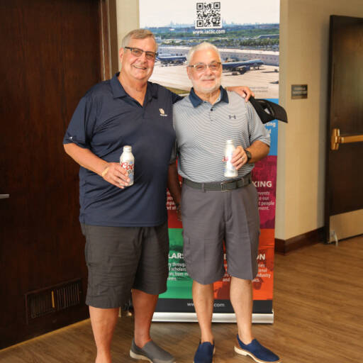 Two men are standing side by side, each holding a beverage can, and smiling at the camera. Behind them, a retractable banner of the International Air Cargo Association of Chicago (IACAC) announces the 2021 Golf Outing. Both are dressed in casual attire.