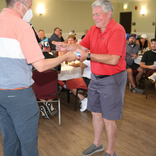 Two men exchanging an item in a room with seated people in the background. The man on the left, wearing a mask, red and gray shirt, and dark pants, hands it over. The man on the right, in a red polo and shorts, smiles as he accepts it during the 2021 IACAC Golf Outing.