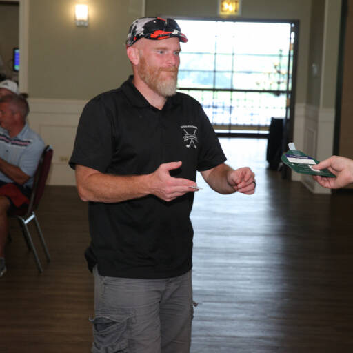 A man with a beard wearing a camo cap, black polo shirt, and gray shorts is extending his hand to receive an item from an unseen person during the 2021 IACAC Golf Outing. He is standing in a room with wooden flooring and people sitting in the background.