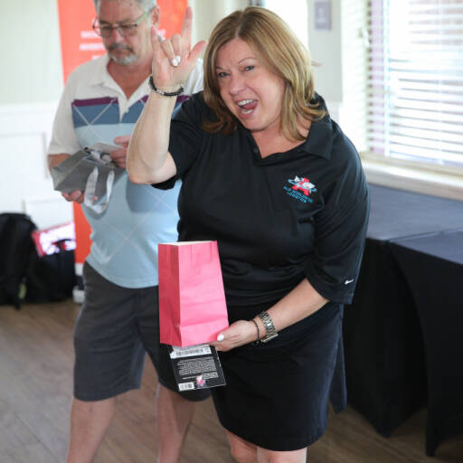 A woman in a black polo shirt holds up a pink gift bag with a playful expression and gestures with her hand. Behind her, a man with gray hair and glasses looks down at a white object he's holding. They are both indoors, likely at the IACAC 2021 Golf Outing, with a red and white banner partially visible in the background.