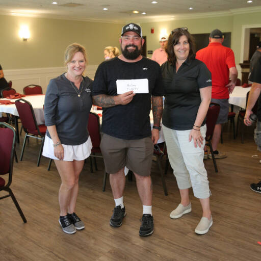 A group of three people stand indoors in a room with red chairs and white table covers. The person in the middle, possibly back from a Golf Outing, holds an envelope and wears a black cap, black shirt, and shorts. The two individuals on either side wear casual attire and smile towards the camera, embodying the IACAC spirit.