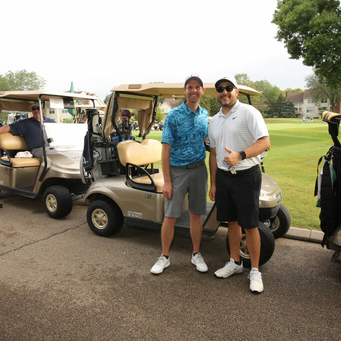 A group of men stands near golf carts on a paved path at the IACAC Golf Outing 2021. Two men in the foreground are smiling and posing for the photo, while two other men load golf clubs into the carts in the background. Trees and houses are visible in the distance.