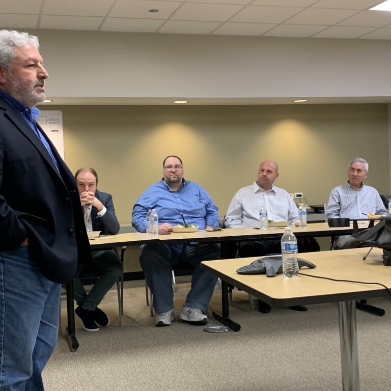 A man with gray hair and a beard is standing and speaking in a conference room during an April 2019 luncheon. Four seated men are attentively listening to him. A projector and water bottles are on the table in front of them. The room has white walls, a beige floor, and ceiling lights.