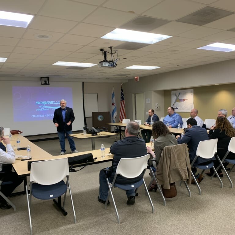 A man stands in front of a large screen giving a presentation during an April 2019 luncheon, to a group of people seated around tables in a conference room. The slide on the screen reads "Syscoel Systems: Delivering New Opportunities." American and state flags are visible in the background.