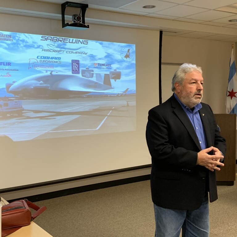 A man in a dark blazer presents in front of a projection screen displaying a presentation slide about Sabrewing Aircraft Company during an April 2019 luncheon. The slide includes images of a drone and logos of various sponsors. Flags of the United States and Chicago are visible in the background.