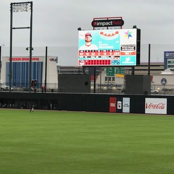 A baseball field featuring an outfield with a large digital scoreboard displaying player information and stats. The background includes stadium advertisements and buildings, set against a cloudy sky overhead. Several players can be seen in the outfield during the IACAC 2018 Baseball Networking Event.