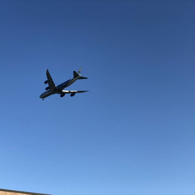 A large airplane with four engines is flying in a clear blue sky, seemingly approaching for landing with landing gear extended. The image's bottom edge shows part of a building or structure, perhaps preparing for the bustling events like the 2018 IACAC or September Oktoberfest.