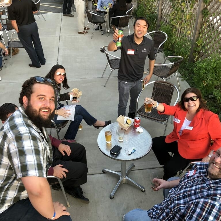 A group of five people sit and stand around a small outdoor table, smiling and holding drinks. The area features a lattice fence and greenery in the background, reminiscent of a lively Oktoberfest gathering. Nearby tables are also filled with cheerful guests enjoying the September festivities.