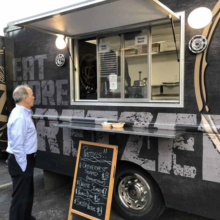 A man stands in front of a food truck with an open window, displaying the menu and prices for various items including pretzels. The truck exterior is decorated with graphics and the text "Eat More Pretzels." Celebrating Oktoberfest, a chalkboard at the front lists menu items and prices.