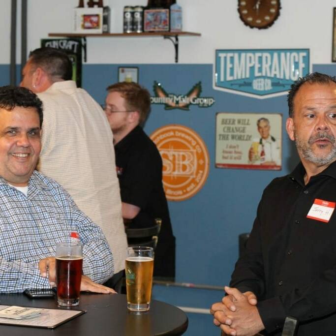 Two men are standing at a high table with drinks in a casual indoor setting, reminiscent of an Oktoberfest gathering. The man on the left is smiling and wearing a checkered shirt, while the man on the right, sporting a black shirt and 2018 IACAC name tag, looks off to the side. Various signs and posters adorn the wall behind them.