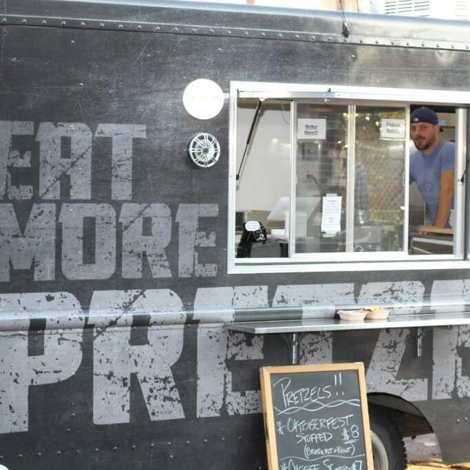 A food truck painted with the words "EAT MORE PRETZEL" in large letters is ready for business. The side window is open, revealing a person in a blue beanie and grey shirt. A chalkboard menu outside promotes pretzels and an Oktoberfest special for $8, celebrating September 2018 IACAC.