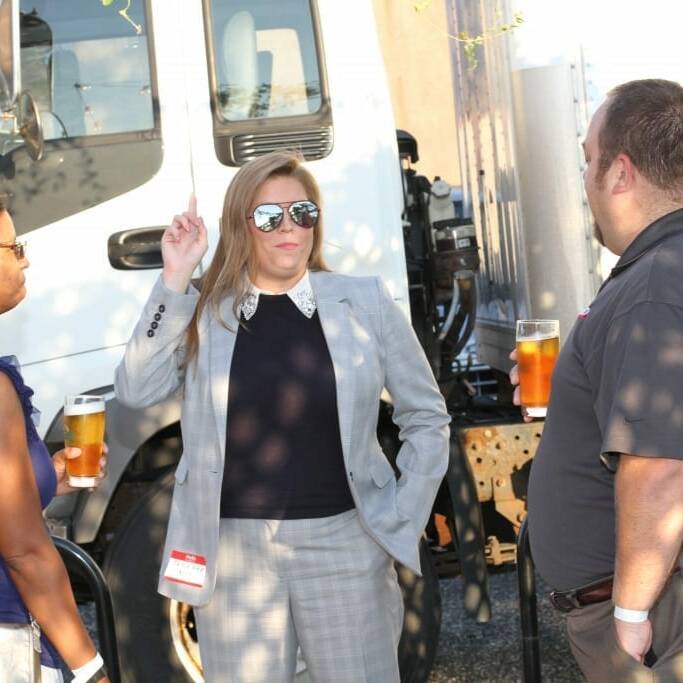 Three people are gathered outdoors near a truck during an Oktoberfest event. The person in the middle, wearing a grey suit and sunglasses, holds a finger up while speaking. The person on the left, in a blue top, and the person on the right, in a dark shirt, both hold drinks in their hands.