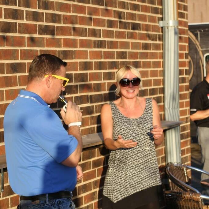 Two people are talking and laughing in an outdoor setting near a brick wall at what appears to be an IACAC 2018 Oktoberfest event. The man on the left is wearing a blue shirt and yellow sunglasses, holding a beer. The woman on the right is wearing a patterned top and large black sunglasses. Another man is in the background.