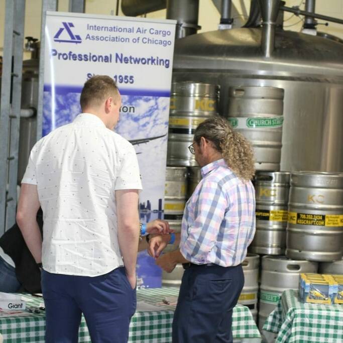 Two men are having a conversation next to an exhibition table at a professional networking event, with the "International Air Cargo Association of Chicago" banner displayed prominently in the background. The setting appears to be an industrial space with large stainless steel tanks and kegs, reminiscent of the 2018 IACAC September Oktoberfest.