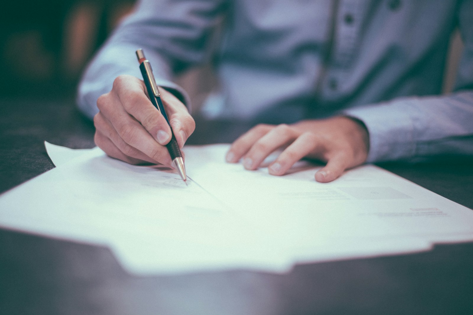 A person in a blue shirt writes on a piece of paper with a pen. Several sheets of paper, including the President's Letter from April 2020, are spread out on the table. The focus is on the hands and the pen, while the rest of the scene is slightly blurred.