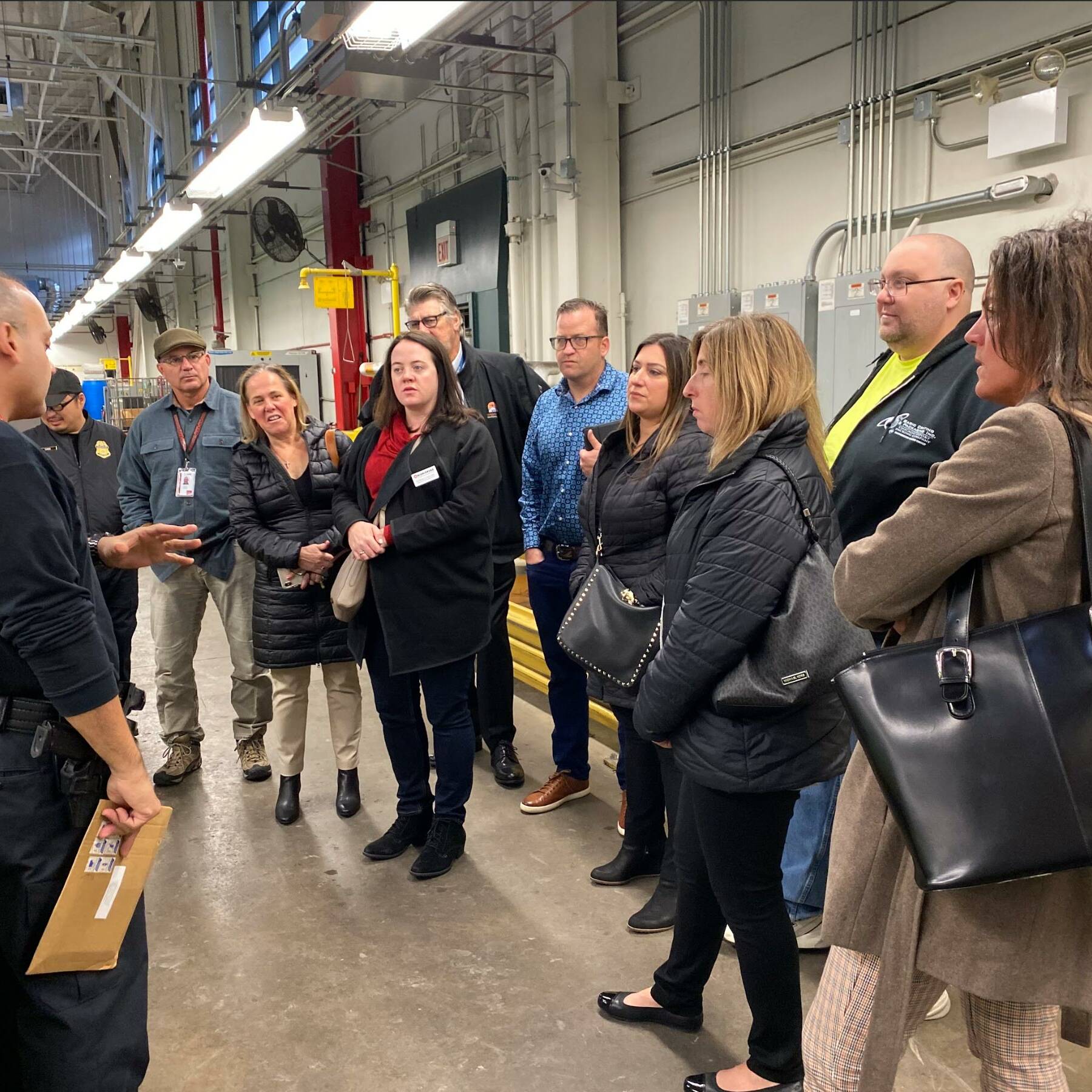 A group of people stand in a semi-circle inside an industrial setting, attentively listening to an individual in a black shirt with 