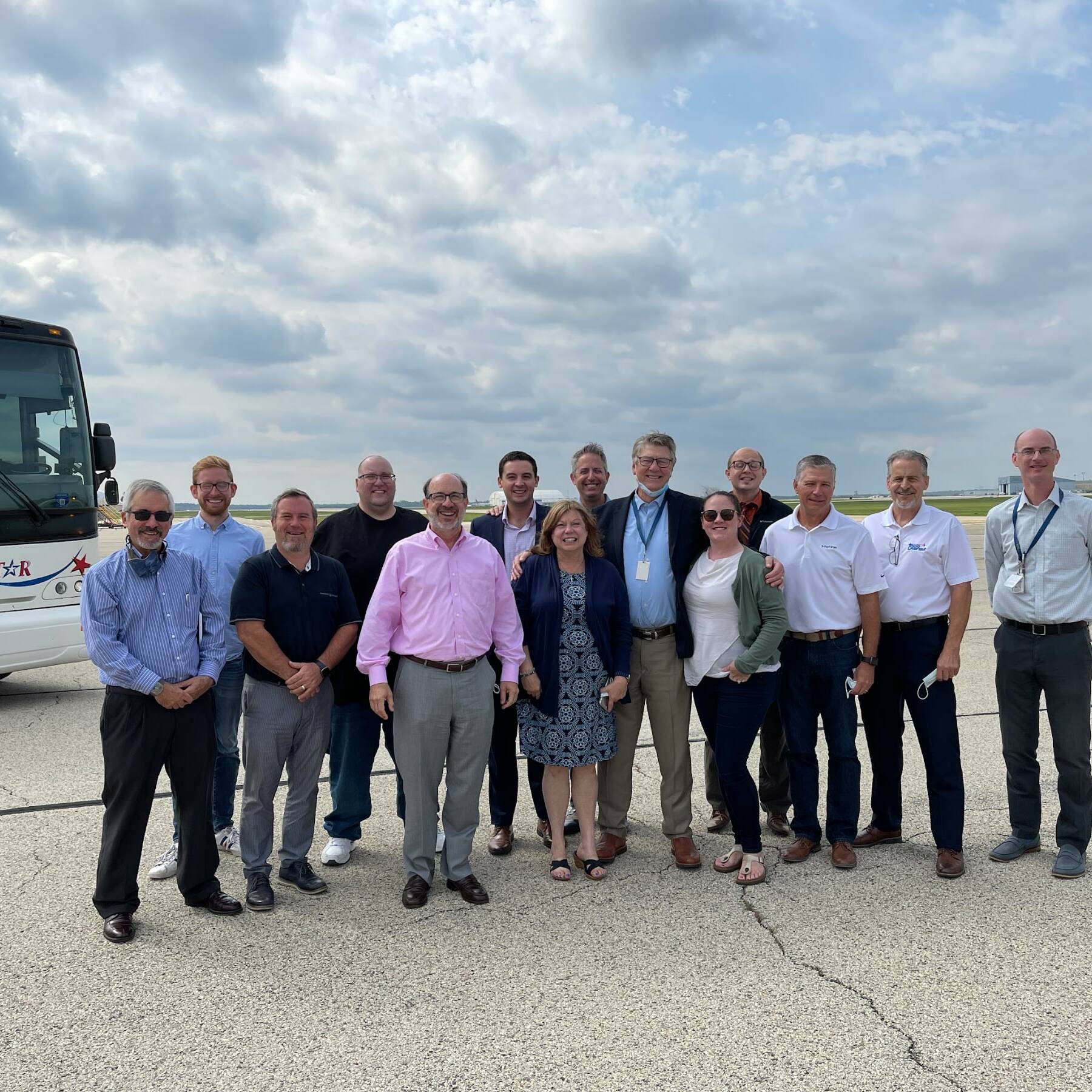 A group of people, including men and women, stands together and smiles for a photo on an airport tarmac. Behind them is a white bus with 